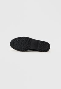 Fekete sneaker talp, texturált mintázattal, kör alakú tapadóelemekkel és kiemelkedő vonalakkal a tapadás érdekében. A márkajelzés látható az oldalon.