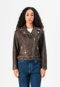Brun skinnmotorsykkeldress med belte i midjen, glidelåslommer og sølvfarget utstyr. Har notert krage og teksturert overflate.
