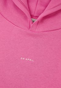 Rozā kapuce ar mīkstu tekstūru, ar auklu regulējamu kapuci un baltu tekstu uz priežas: "OH APRIL" priekšpusē. Skats no augšas.