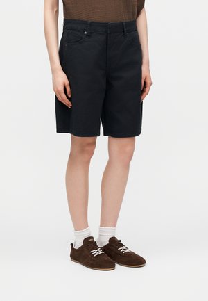 Άτομο που φοράει μαύρα σορτς μέχρι το γόνατο, καφέ αθλητικά παπούτσια με λευκά κορδόνια, λευκές κάλτσες και καφέ τοπ, στέκεται μπροστά σε ένα απλό φόντο.