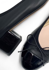 Ballerine nere con punta arrotondata, tomaia in pelle testurizzata, dettagli in vernice lucida e tacco largo basso. Presenta un fiocco decorativo.