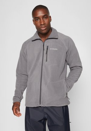 Chaqueta de forro polar gris con cierre frontal, cuello alto y dos bolsillos laterales. Presenta un logotipo en el pecho y una textura suave y lisa.