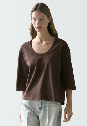 Jonge vrouw die een loszittende bruine top met een scoop-hals en ellebooglange mouwen draagt, en lichtblauwe verweerde spijkerbroek, staand tegen een effen achtergrond.