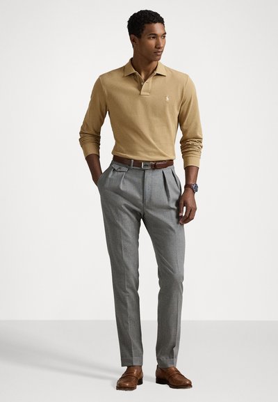 Bēžs garām piedurknēm polo krekls, pelēki plisēti bikses, brūns ādas jostas, un brūni ādas apavi, ar rokaspulksteni uz kreisās rokas.