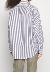 Persona con capelli ricci che indossa una camicia bianca a maniche lunghe, larga, con strisce verticali nere e pantaloni beige, mostrata di spalle.