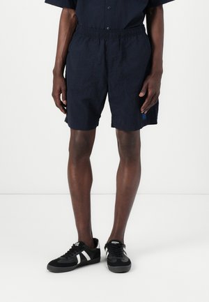 Homme portant un short noir à taille élastique et des baskets noires avec des bandes blanches, debout devant un fond blanc uni.