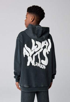 Jeune personne portant un sweat à capuche noir ample avec le texte blanc "ALLSAINTS LONDON" dans le dos, debout devant un fond gris uni.