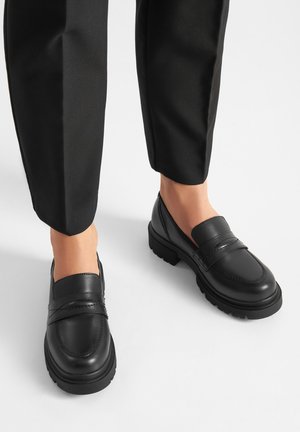 Czarne skórzane mokasyny z zaokrąglonym noskiem, przeszyciami i niską platformą; zestawione ze zwężanymi czarnymi spodniami na białym tle.