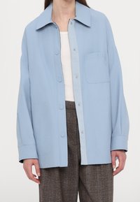 Světlomodrá košilová bunda s límečkem a našitou kapsou. Vyrobená z hladké látky, má volný střih a jemný detail loga.