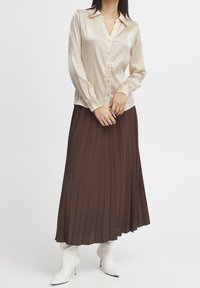 Chemise en soie beige à boutons avec des manches longues, associée à une longue jupe plissée marron foncé. Le mannequin porte des bottes blanches à talons.