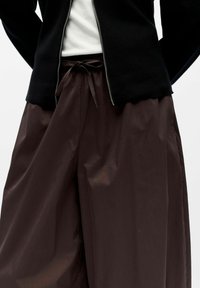 Veste zippée noire, superposée sur une chemise blanche ; pantalon marron ample avec des liens à la taille. Le tissu semble lisse et légèrement brillant.