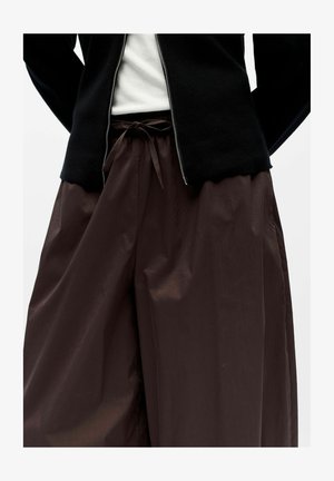 Crna jakna na patent zatvarač, složena preko bijele košulje; široke smeđe hlače svezane oko struka. Tkanina izgleda glatko i pomalo sjajno.