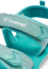 Tyrkysové sandály se třpytivými popruhy, texturovanou stélkou a tvarovanou podrážkou. Obsahují logo Hummel a nastavitelné zapínání.