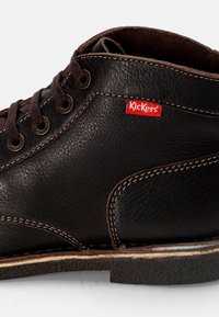 Stivale in pelle nera alla caviglia con lacci marroni, dotato di occhielli metallici, un logo Kickers rosso ben visibile e cuciture bianche a contrasto lungo i bordi.