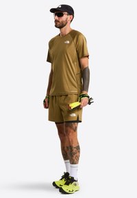 Olive green athletic shirt and shorts with black accents, paired with neon yellow running shoes. Accessories include a black cap and sunglasses.