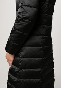 Schwarzer, gepuffter Mantel mit gesteppter Oberfläche, der eine weiche, glänzende Textur und lange Ärmel hat, von der Seite ohne Kapuze betrachtet.