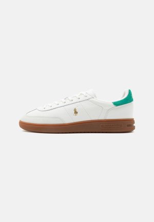 Fehér alacsony szárú sneaker barna talppal, zöld sarokfüllel és arany Polo-logóval az oldalon, egyszínű háttér előtt.