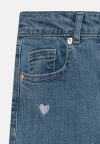 Traperice u srednje plavoj boji s malim svijetloplavim srcem izvezеним na prednjoj džepu, klasičnog dizajna i metalnim gumbom.