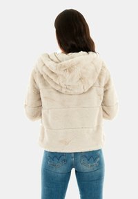 Chaqueta con capucha de piel sintética en color crema, con una textura suave y costuras horizontales. Combinada con unos vaqueros ajustados de color azul claro.
