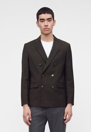 Young man wearing a dark brown double-breasted blazer with subtle plaid pattern and grey trousers, standing against a plain background.