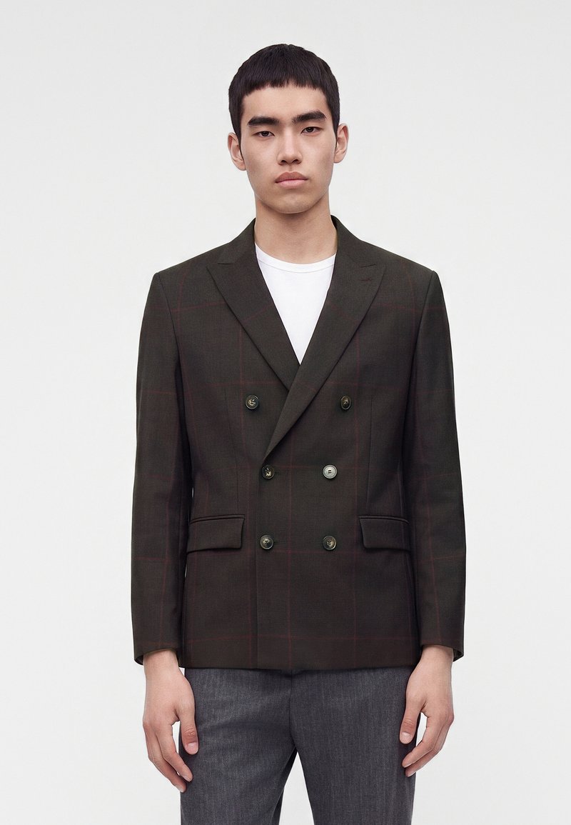 Jeune homme portant un blazer croisé marron foncé avec un motif à carreaux subtil et un pantalon gris, debout devant un fond uni.