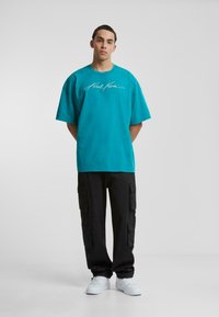 Tirkīza krāsas pārāk liels T-krekls ar baltu parakstu, kombinēts ar melniem cargo biksēm, kas aprīkotas ar vairākiem kabatām, un baltām kedām.