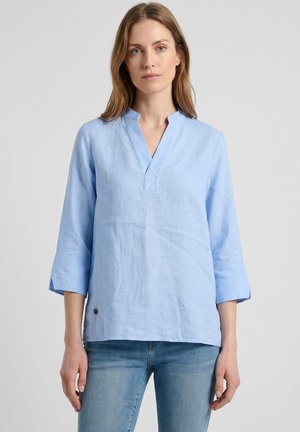 Mujer con una túnica de lino azul claro con cuello en V partido y mangas tres cuartos, combinada con vaqueros de mezclilla azul claro.