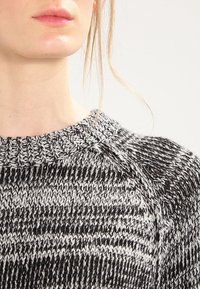 Zbliżenie na osobę w czarno-białym swetrze z melanżowej przędzy, z okrągłym dekoltem z prążkowanym wykończeniem i luźnymi pasmami blond włosów.