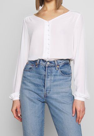 Mujer que lleva una blusa blanca de manga larga con botones forrados de tela y vaqueros azul claro de talle alto sobre un fondo liso.