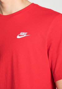 Červené tričko Nike s vyšívaným bílým logem Nike na levé hrudi, nošené osobou viditelnou od krku po horní část paže.