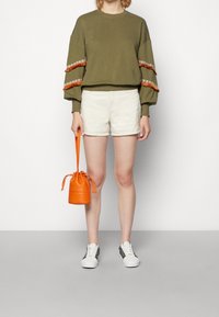 Felpa verde oliva con dettagli a frange arancioni, pantaloni corti bianchi e una piccola borsa in pelle arancione. Sneakers bianche con lati decorati.
