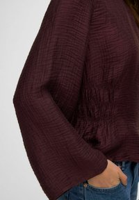 Vinrød, tekstureret bluse med brede ærmer og detaljeret, smocket talje. Kombineret med blå denimjeans med sidelomme.