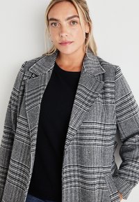 Σακάκι καρό σε λευκό και μαύρο με πέτο, υφή υφάσματος και λεπτομέρεια με κουμπιά μπροστά, στρωμένο πάνω από ένα απλό μαύρο μπλουζάκι.