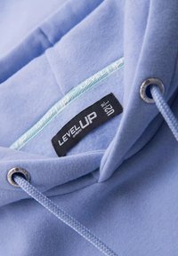 Haut à capuche bleu clair avec une texture douce, cordon de serrage avec œillets en métal, et une étiquette noire portant la mention "LEVEL UP". L'intérieur présente une bordure à motifs.