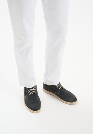 Czarne perforowane buty z beżowymi podeszwami i sznurowadłami, noszone z białymi spodniami. Buty mają gładką teksturę i casualowy design.