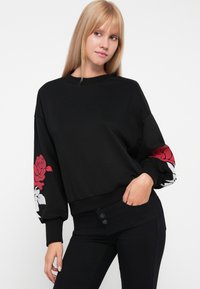 Sudadera negra con mangas globo que presenta diseños florales en rojo y blanco en los puños. El material es suave, con una cinturilla ajustada.