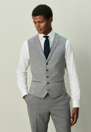 Mand iført grå jakkesætvest og bukser, hvid skjorte og mørkt slips, står mod en neutral baggrund med hænderne i lommerne.