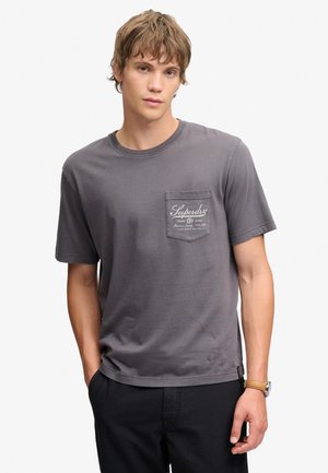 Ung mand iført en grå Superdry t-shirt med brystlomme og sorte bukser, stående med den ene hånd i lommen mod en ensfarvet baggrund.