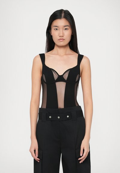 Woman with long dark hair wearing a black sleeveless mesh corset top and high-waisted black pants with a wide belt and silver buttons.
