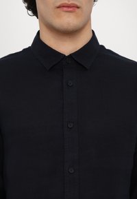 Zwarte button-up shirt van gladde stof, met een kraag en contrasterende knopen; subtiele logo-detail op de linkerborst.