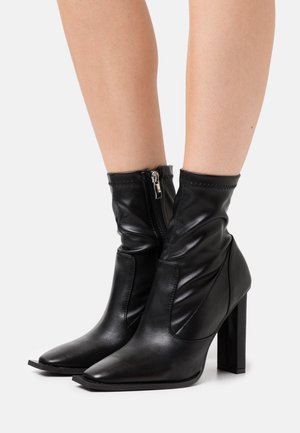 Bottes noires en cuir à cheville avec une pointe pointue et un talon bloc, présentant une fermeture éclair sur le côté et une texture froncée en haut.