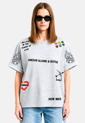 Femme aux cheveux bruns raides portant des lunettes de soleil noires et un t-shirt gris avec différents imprimés graphiques noirs et colorés ainsi qu’un texte.