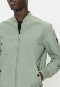 Világoszöld bomber dzseki cipzáros elülső résszel, bordázott mandzsettákkal és két oldalzsebbel. Simább, könnyű anyag, visszafogott dizájn.