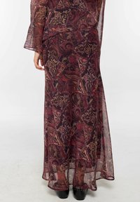 Mujer vistiendo una larga falda transparente con un patrón de paisley rojo y negro, combinada con botas de tobillo negras con tachuelas, de pie sobre un suelo blanco.