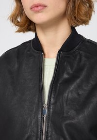 Svart läder bomberjacka med ribbad krage, försedd med en silverfärgad dragkedja och en texturerad grön tröja under.