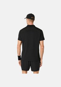 Uomo con cappellino nero, maglietta nera a maniche corte, pantaloni corti neri e bracciale nero, in piedi di spalle contro uno sfondo neutro.