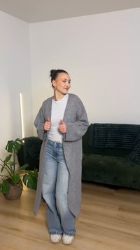 Pelēks, garš kardigans ar vaļēju piegriezumu un platām piedurknēm, nēsāts pār gaiši pelēku augšu un zilām džinsām, attēlots modernā interjerā.