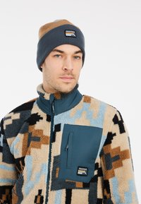 Fleece jakna s mnoštvom geometrijskih uzoraka u bež, plavoj i crnoj boji; ima patentni zatvarač na prsnom džepu i visoki ovratnik.
