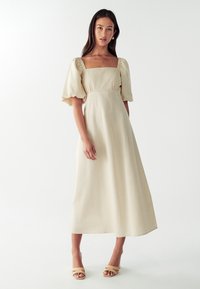Bež haljina od lana s pufastim rukavima, kvadratnim izrezom i strukiranim strukom. Polu-dužine s glatkom teksturom. Uključuje sandale s trakama.