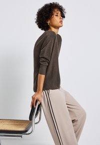 Maglione marrone scuro con polsini a coste, abbinato a pantaloni beige chiaro con due strisce laterali. Il modello tiene in mano una sedia con seduta intrecciata.